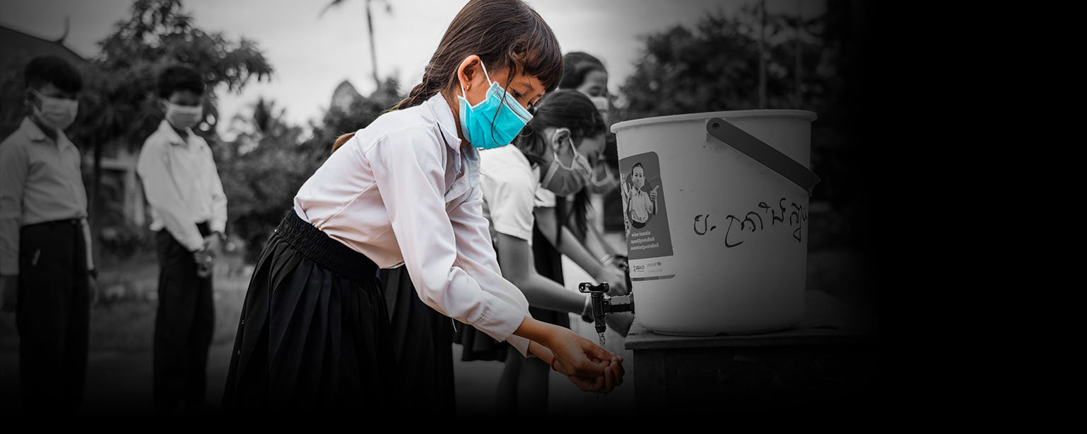 a girl washes her hands outside at a public water bucket dispenser with other children, all of whom wear face masks