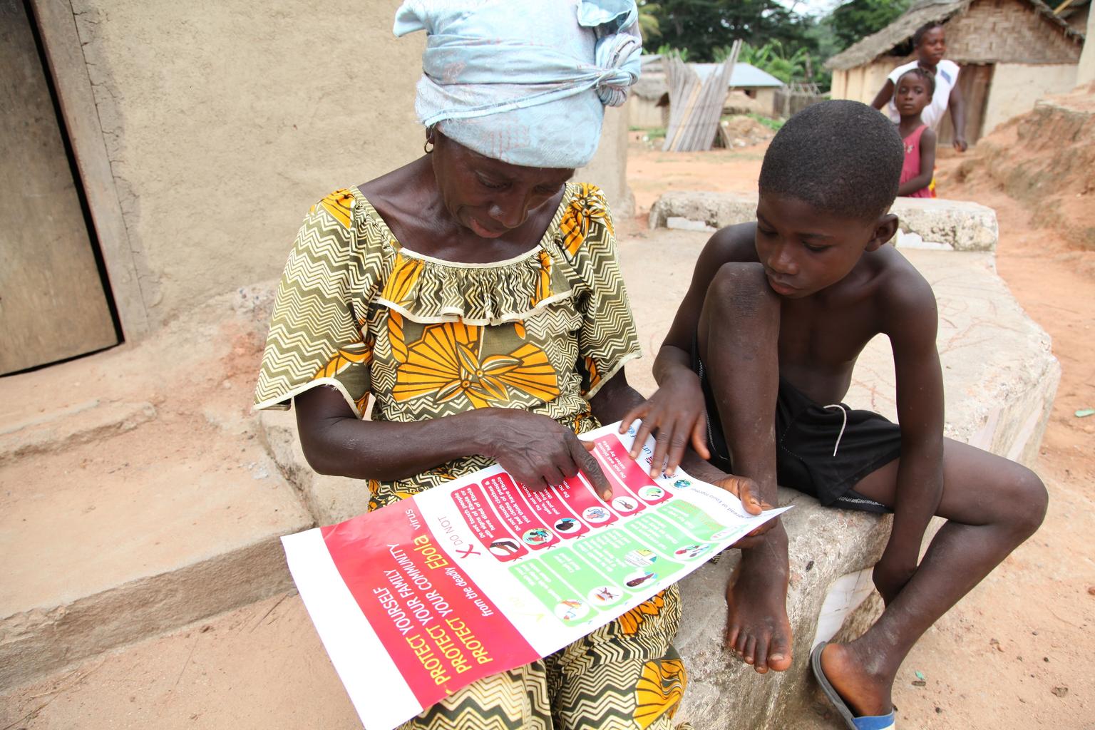 A woman speaks with a young boy in her community about the symptoms of Ebola virus disease (EVD) and best practices to help prevent its spread, in the city of Voinjama, in Lofa County. While speaking, she is referencing a poster, distributed by UNICEF, th