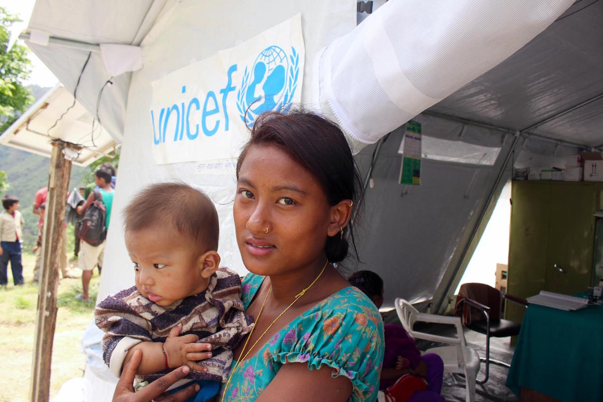 A young woman holds her newborn at the entrance to a tented, makeshift health post provided by UNICEF in the village development committee of Thuladurlung, in Lalitpur District. One of the tent’s tarpaulins bears the UNICEF logo.