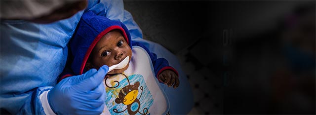 An infant gets spoon fed by a person wearing PPE