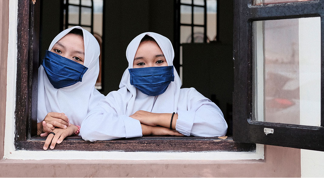 two girls in headwraps and face masks lean on an open window frame looking out