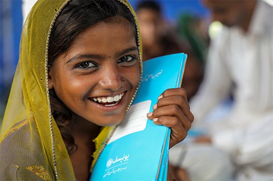 A girl in a headscarf smiles while holding a blue notebook to the side of her face