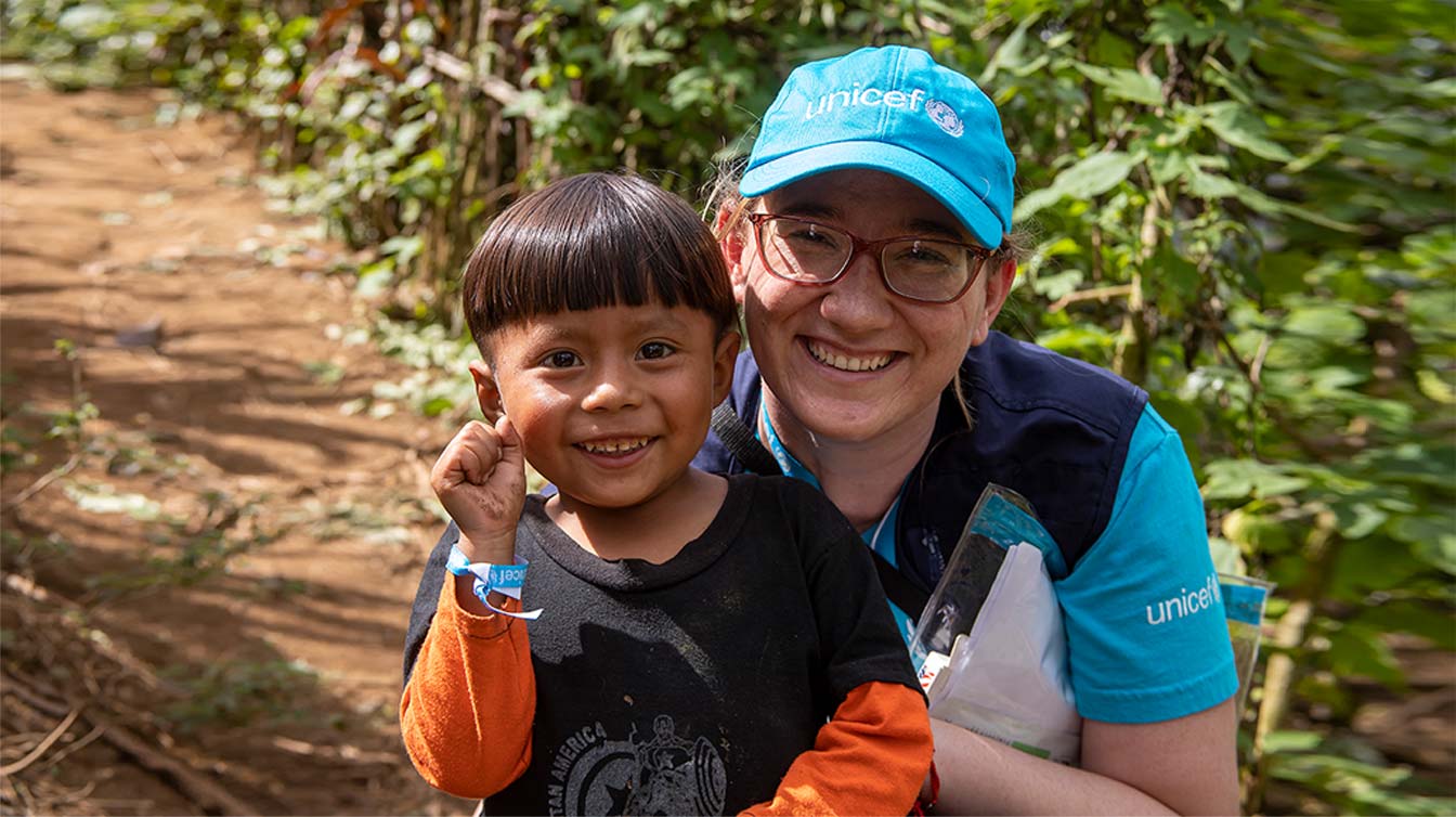 A child and aid worker smile at the camera standing together in a field