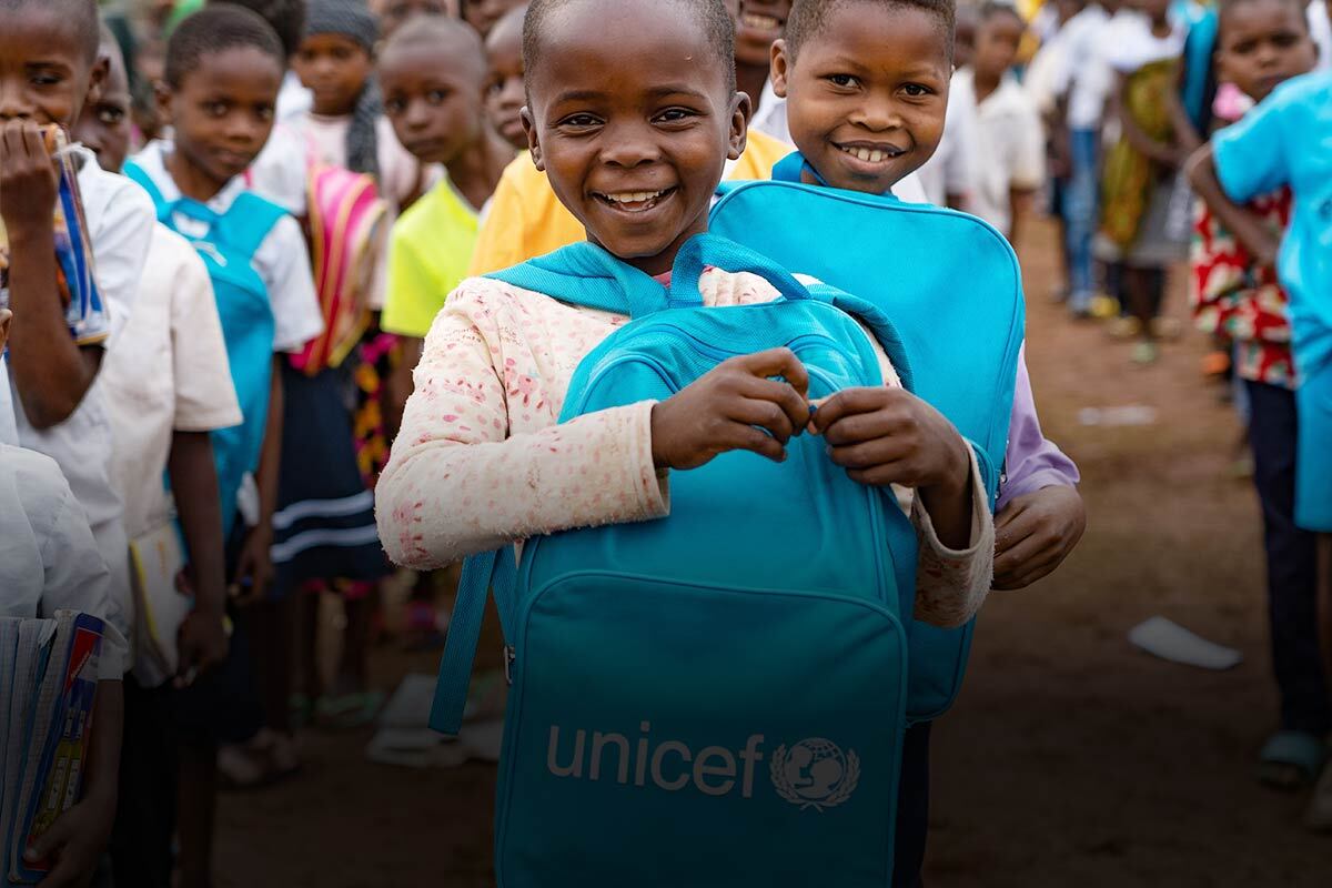 A girl standing in front of other children smiles while holding a blue UNICEF backpack to her chest.