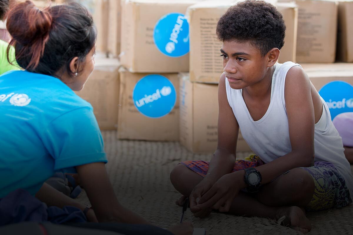 UNICEF workers talk to a boy as they all sit in front of UNICEF supply boxes