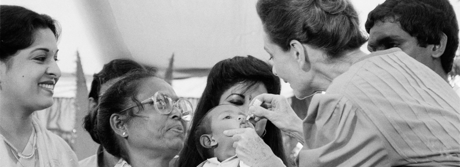 Surrounded by other women and a male health worker, UNICEF Goodwill Ambassador Audrey Hepburn vaccinates a toddler against polio, at a health clinic.