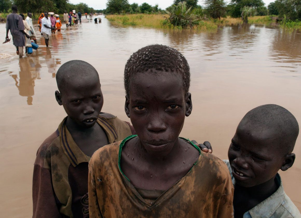 Boys stand in waters caused by seasonal flooding, on a road in South Sudan. Behind them, other pedestrians wade through the water. The county is home to four camps for Sudanese refugees.
