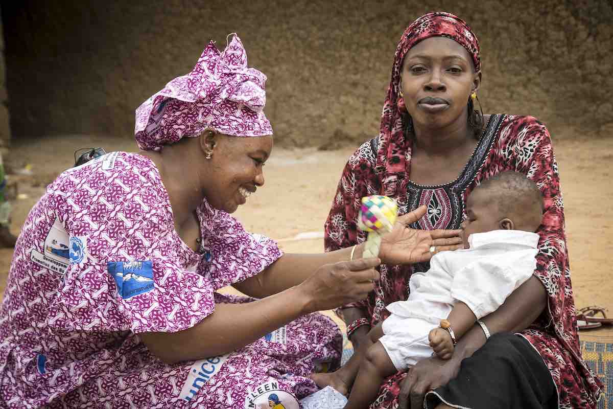 Mother Djénéba Diarra holds Assitan Doumbia, 3 months, as (left) Mama Yeleen Fatoumata Ouattara shakes a rattler to encourage play, during her daily door-to-door visit of families with young children in Baraouéli village, Ségou Region, Mali