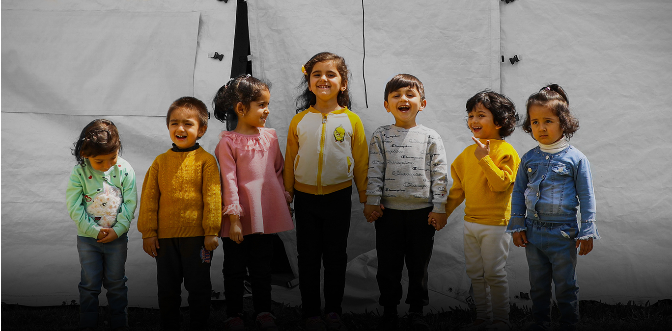 A group of children stand with various expressions on their faces in front of a large tent 