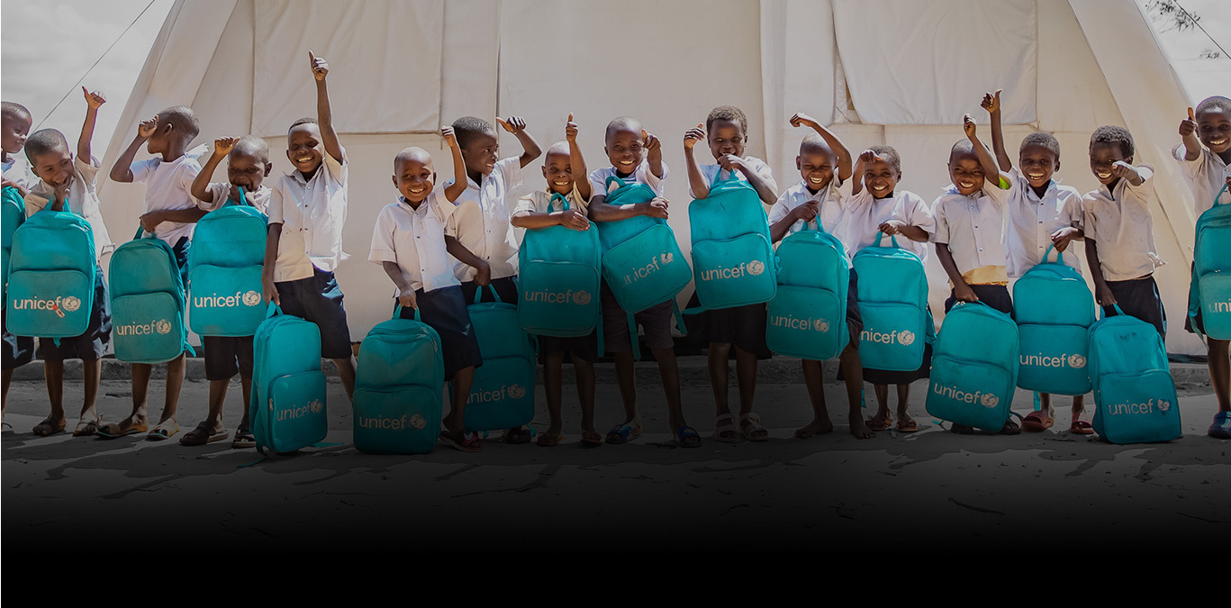 A group of children in front of a large tent hold up their blue UNICEF backpacks