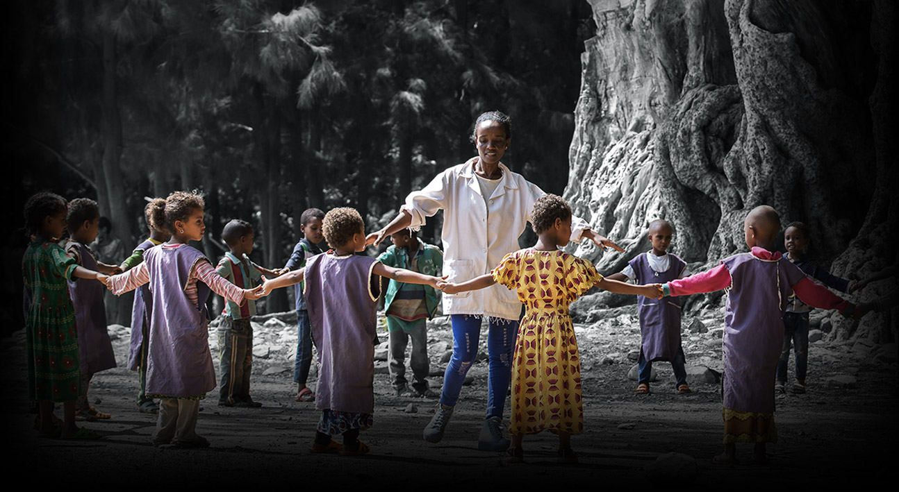 A woman holds hands with a circle of children. They stand in front of a forest, next to very old and large trees