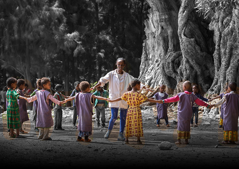 A woman holds hands with a circle of children. They stand in front of a forest, next to very old and large trees