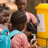 A group of children gather around a water dispenser outside. One child is wearing a blue UNICEF backpack
