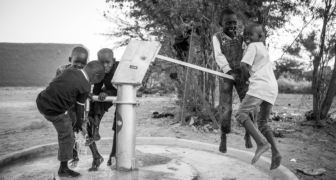 A group of children operate an outdoor water pump together while laughing