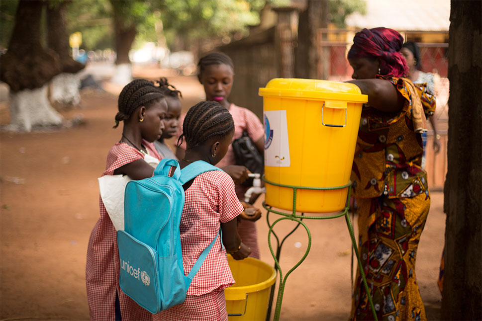 Children get water from an outdoor dispenser. One of the children is wearing the blue UNICEF backpack.