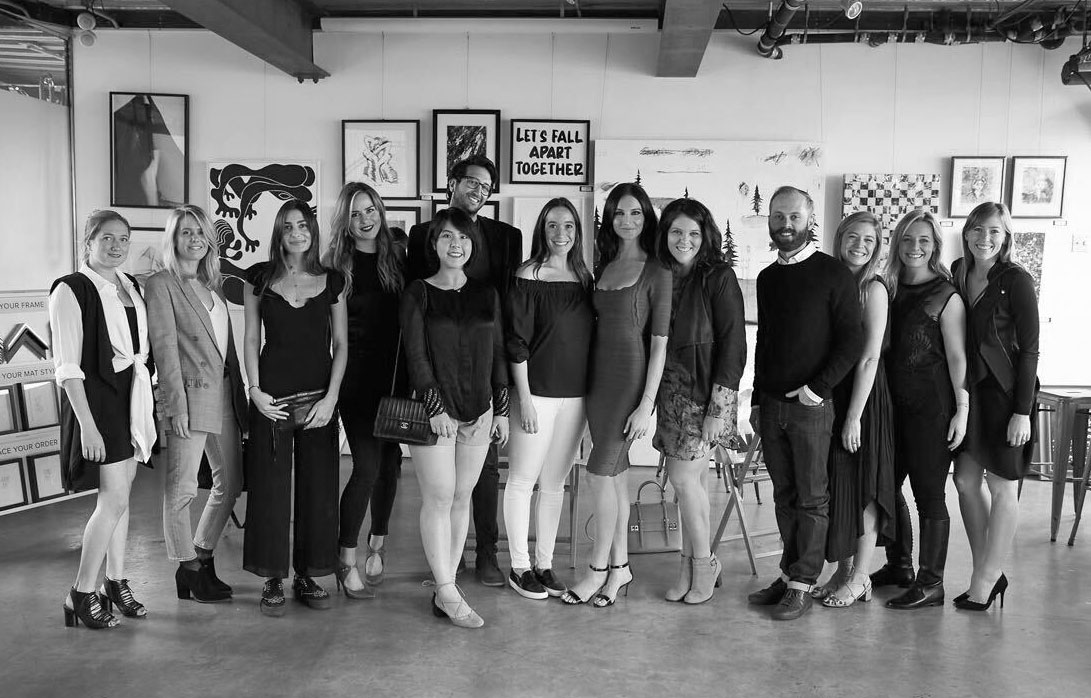 A group of young, professional NextGen members posing at an art gallery. A framed image behind them reads 'let's fall apart together'