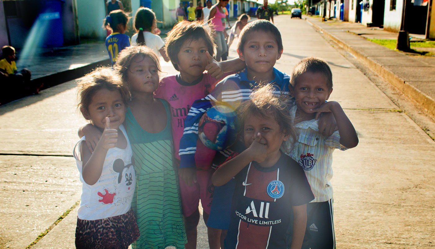 Several children stand on the sreet together with their arms around each other, looking at the camera and smiling. Some make the thumbs up gesture.