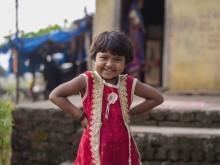 A young girl wearing a red and white dress stands akimbo outside of a house while smiling