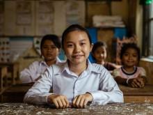 A smiling girl in a dress shirt sits at her desk at school. Other students sit at their desks behind her.