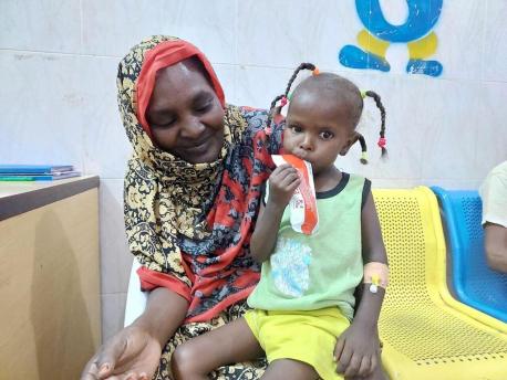 On June 1, 2023, AlBatoul, 3, sits on her mother's lap in Dongola hospital, a UNICEF-supported facility in Sudan's Northern State, working through a packet of nutrient-rich Ready-to-Use Therapeutic Food supplied by UNICEF.