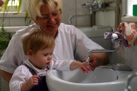 A caregiver shows a toddler child how to wash hands, holding her hand under a sink faucet with the water running.