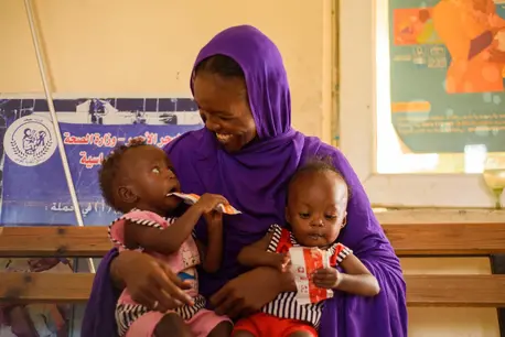 At a UNICEF-supported health facility in Port Sudan, a mother holds her twin daughters in her lap as both consume packets of Ready-to-Use Therapeutic Food, a peanut paste UNICEF supplies to support the recovery of acutely malnourished children.