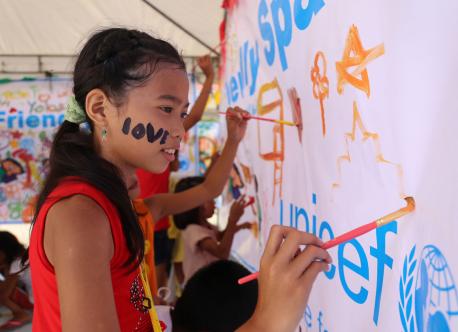 A girl paints a sign with the UNICEF logo in the Philipines. She has love written on her face.