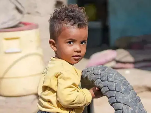 A young child with curly hair stands outside holding a bike tire.
