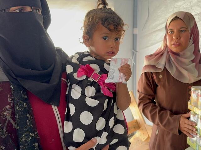 On May 7, 2025, at Al Farooq Camp in Khan Younis, southern Gaza, a woman holds a small child who grips a sachet of Ready-to-Use Therapeutic Food (RUTF).