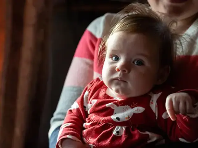 A baby held by her mom, and wearing a red dress with reindeer on them.