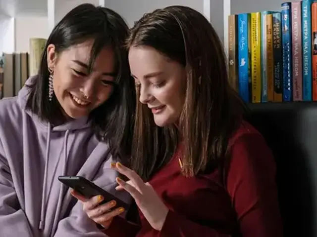 Two girls sitting in front of bookshelves smile looking at a phone together