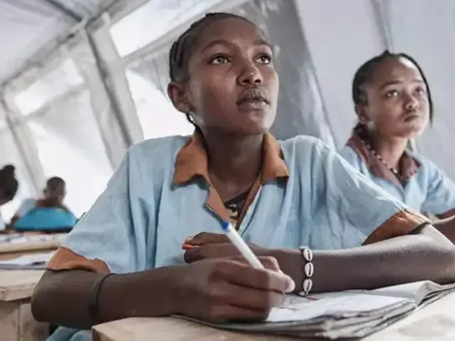 Two girls sit at their desk in front of other students at desks, in a large tent classroom, with their notebooks open, paying attention to a speaker offscreen.