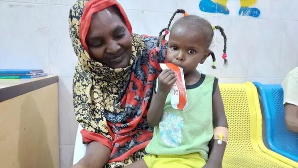 On June 1, 2023, AlBatoul, 3, sits on her mother's lap in Dongola hospital, a UNICEF-supported facility in Sudan's Northern State, working through a packet of nutrient-rich Ready-to-Use Therapeutic Food supplied by UNICEF.