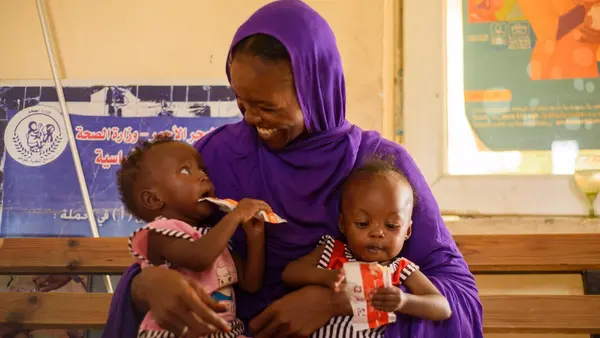 At a UNICEF-supported health facility in Port Sudan, a mother holds her twin daughters in her lap as both consume packets of Ready-to-Use Therapeutic Food, a peanut paste UNICEF supplies to support the recovery of acutely malnourished children.