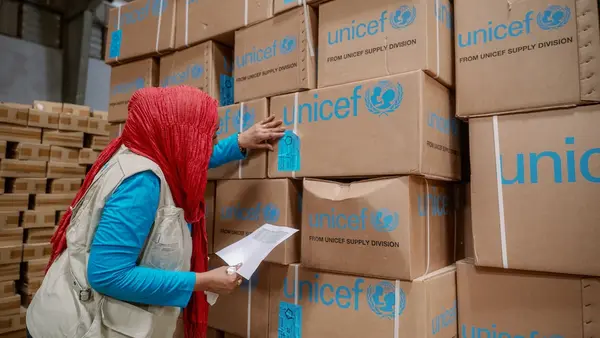 Woman wearing head scarf and vest examines cardboard boxes labeled UNICEF