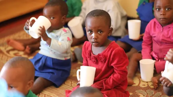Young girl sitting on floor holding white mug looking directly into camera, surrounded by additional children holding and drinking from mugs