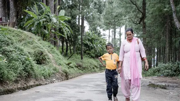 A young boy holding a woman's hand as they walk on a road lined by trees and plants
