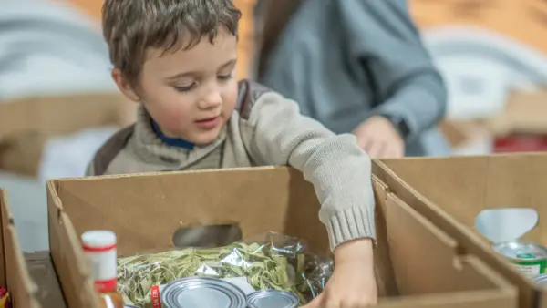 While volunteering with his mother at a local food bank, a young boy smiles and reaches into a cardboard box filled with pantry items.