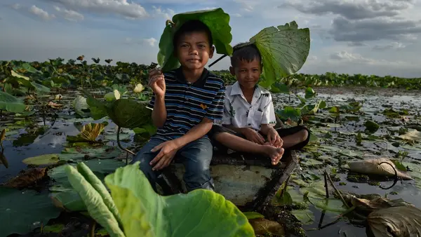 In Thailand during a heat wave in May 2025, two children sit on wet ground holding lotus leaves