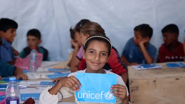 A smiling girl holds a UNICEF notebook in a temporary learning space in the Gaza Strip in 2024.