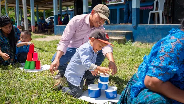 Young boy kneeling and stacking plastic cups while his father kneels behind him and assists