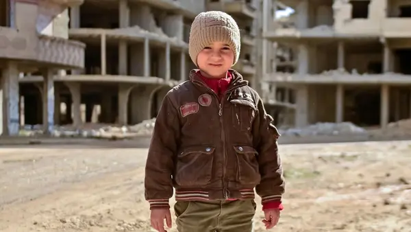 A 5-year-old boy wearing a hat and jacket stands in front of bombed-out buildings in Tadamon, Damascus, Syria on Jan. 14, 2026.