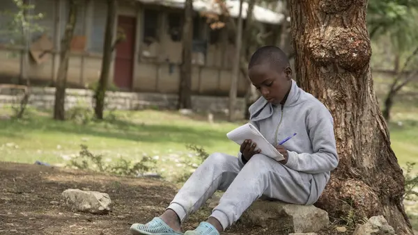 A boy in Ethiopia writes poetry in a notebook sitting by a tree. He uses poetry to help him understand his experiences and feelings.