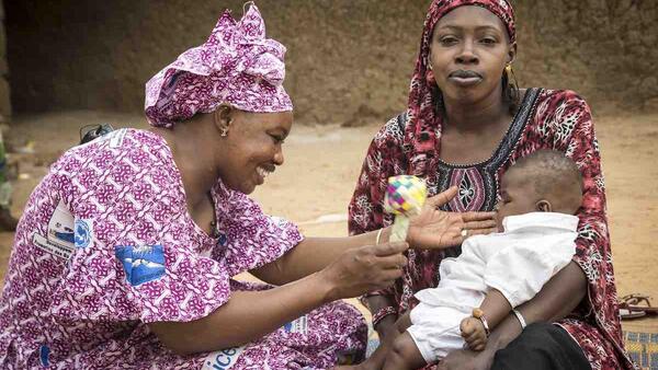 Mother Djénéba Diarra holds Assitan Doumbia, 3 months, as (left) Mama Yeleen Fatoumata Ouattara shakes a rattler to encourage play, during her daily door-to-door visit of families with young children in Baraouéli village, Ségou Region, Mali