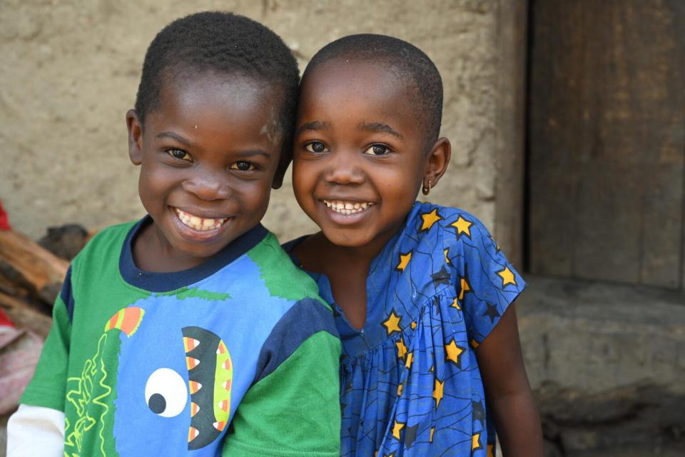 Two children smile brightly at the camera, wearing colorful shirts, sitting outside a building