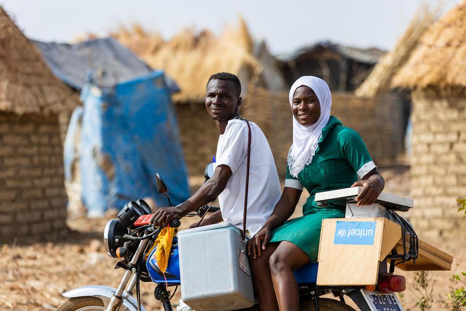 An Immunization Officer and Community Health Nurse supported by UNICEF travel by motorbike to a camp for displaced families in Sapelliga where they will provide services..