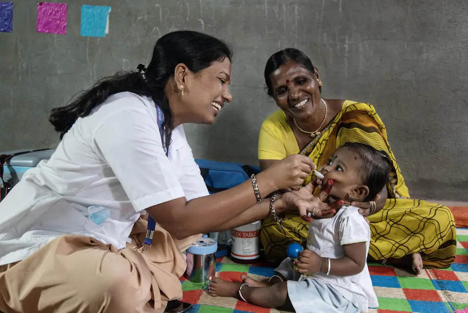 An Auxiliary Nurse Midwife administers a dose of Vitamin A to 9-month-old Jivika with her grandmother in India.