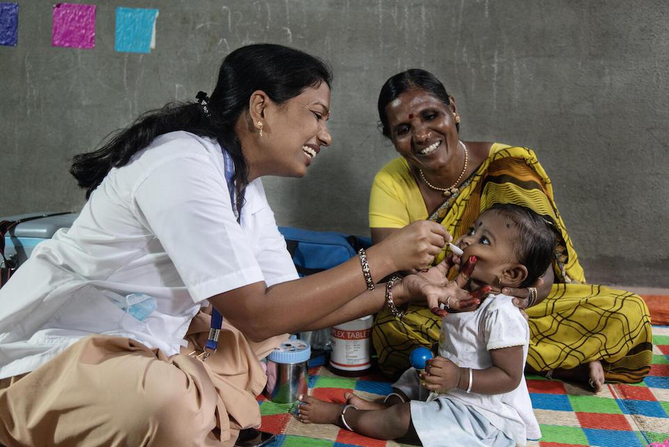 An Auxiliary Nurse Midwife administers a dose of Vitamin A to 9-month-old Jivika with her grandmother in India. 