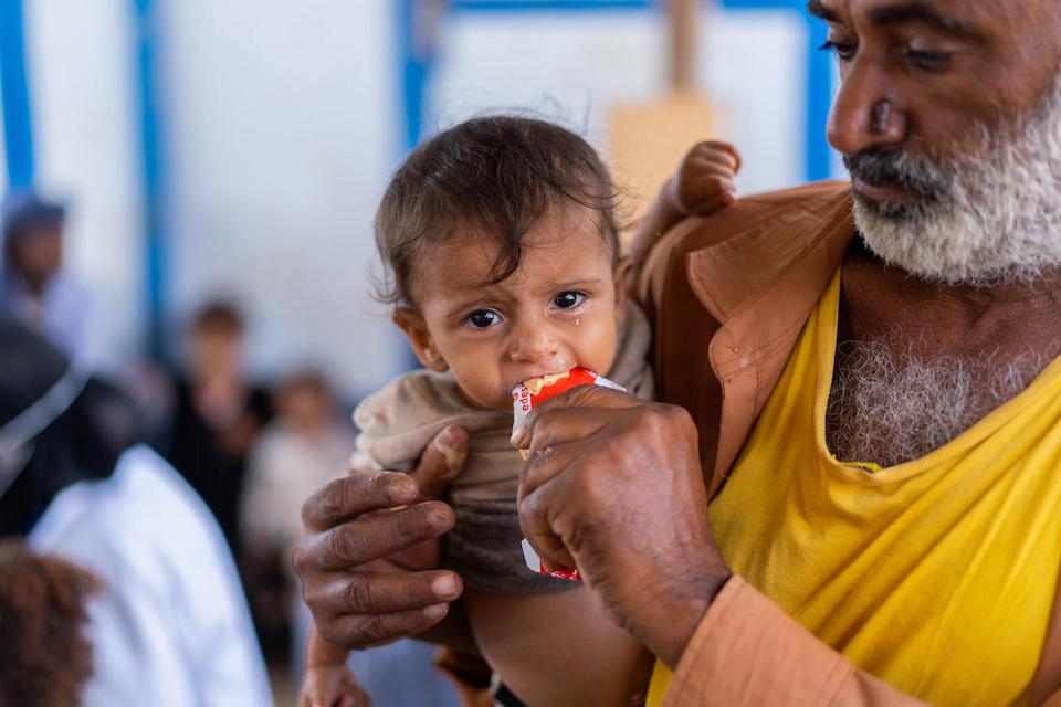 A child eats from a sachet of Ready-to-Use Therapeutic Food (RUTF) while his father holds him in Al-Sha’ab, a camp for displaced families in Al Buraiqeh District, Aden Governorate, Yemen. 