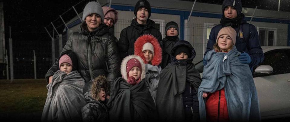A family with children stand wrapped in blankets outside at night in front of a wire fence